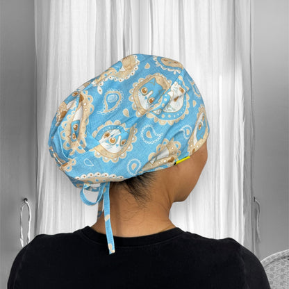 A person wearing a light blue scrub cap adorned with a whimsical pattern of cat faces and decorative swirls.