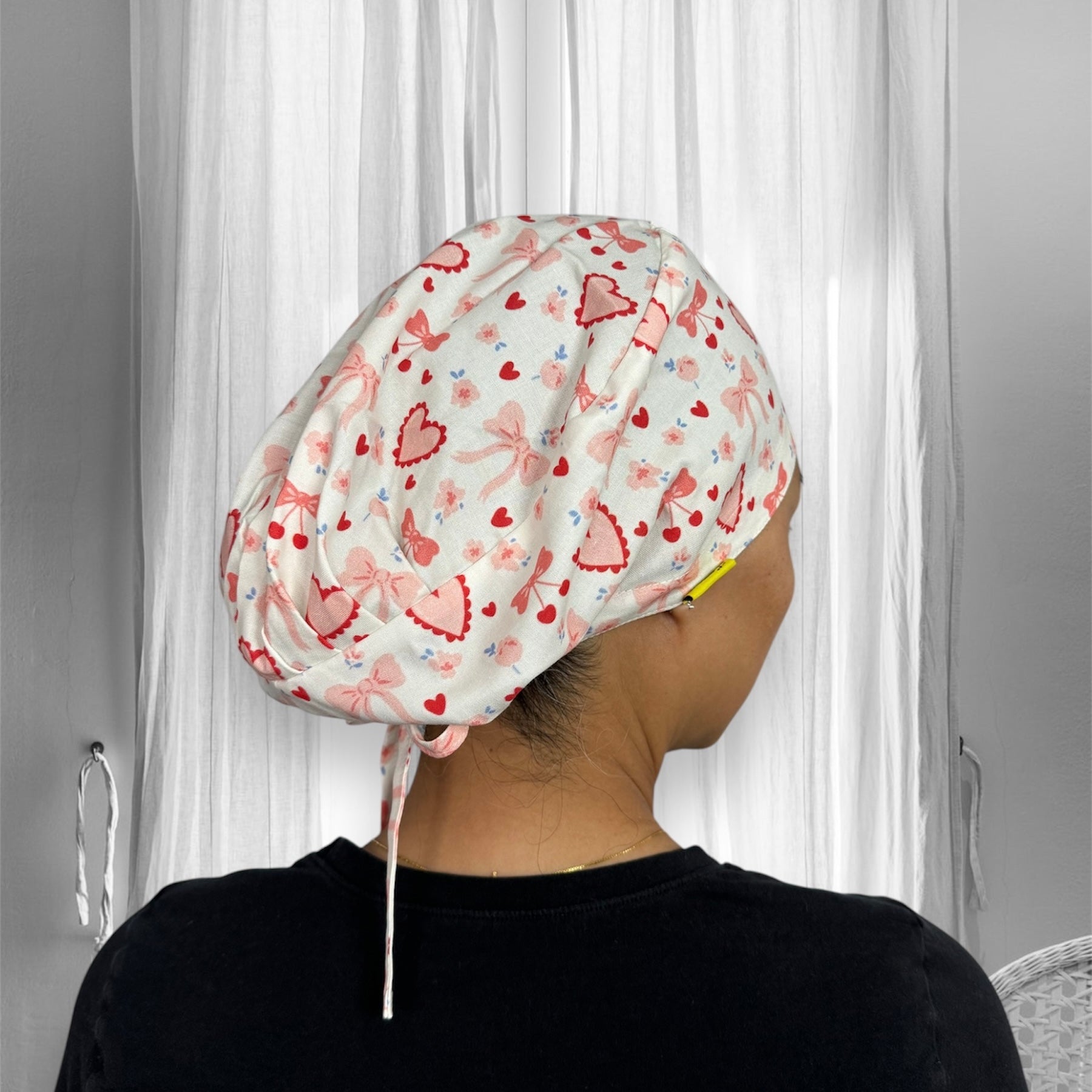 A person is wearing a stylish scrub cap adorned with a cute pattern of pink bows and red hearts.