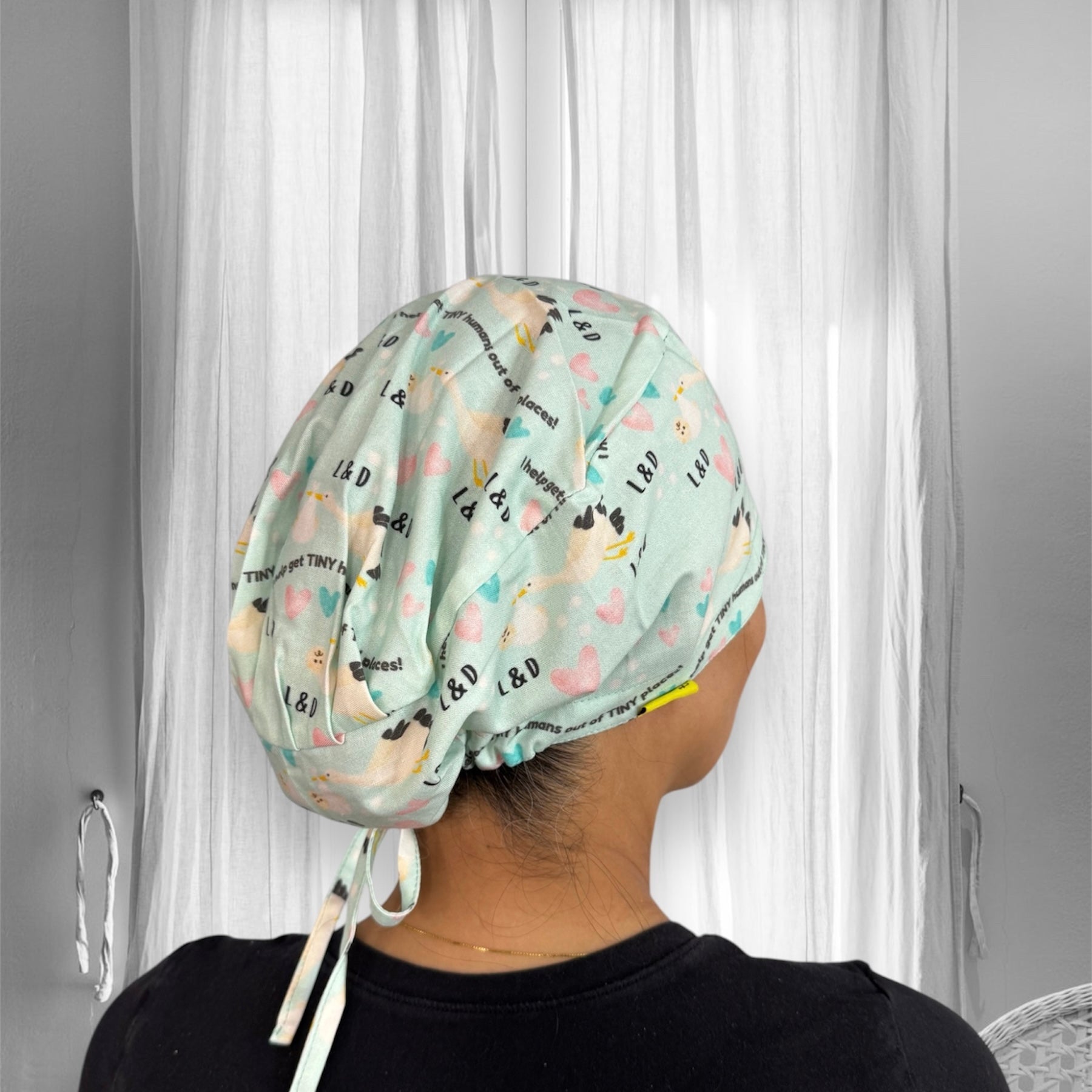 A colorful scrub cap adorned with heart and letter patterns featuring the initials "L & D." The cap is secured with ties at the back and has a whimsical design that includes various small motifs.