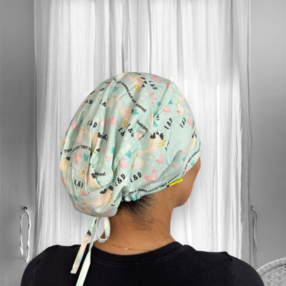 A colorful scrub cap adorned with heart and letter patterns featuring the initials "L & D." The cap is secured with ties at the back and has a whimsical design that includes various small motifs.