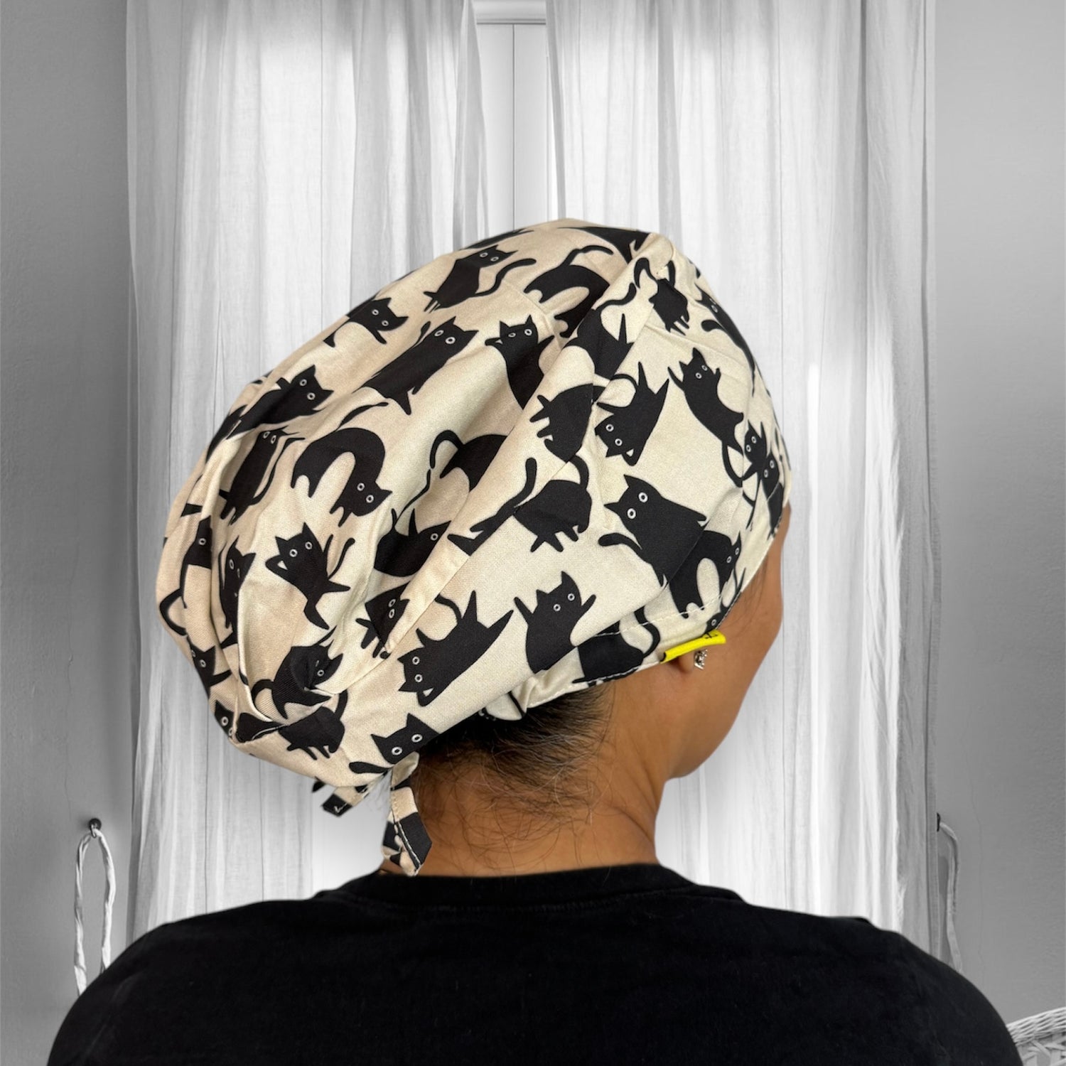 A person wearing a scrub cap with a playful pattern of black cats set against a light background. The fabric is draped elegantly,