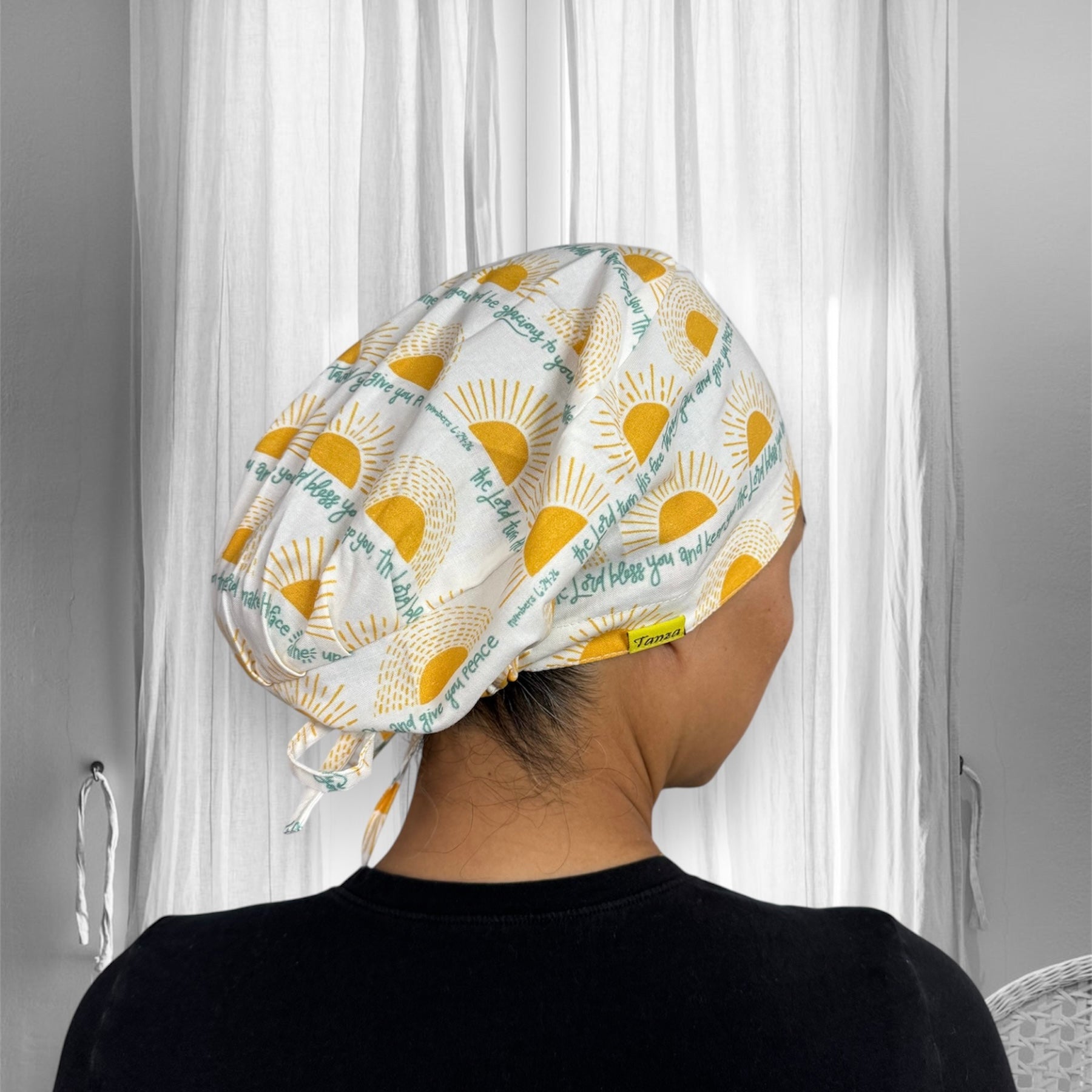 A person wearing a scrub cap with a sun pattern. The cap features yellow suns radiating rays on a light background, interspersed with text phrases in green. The phrases include inspirational or positive messages.
