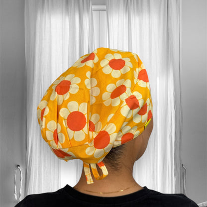 A person wearing a vibrant scrub cap with a retro floral pattern. The cap is predominantly yellow with large white daisies, each having a bright orange center.