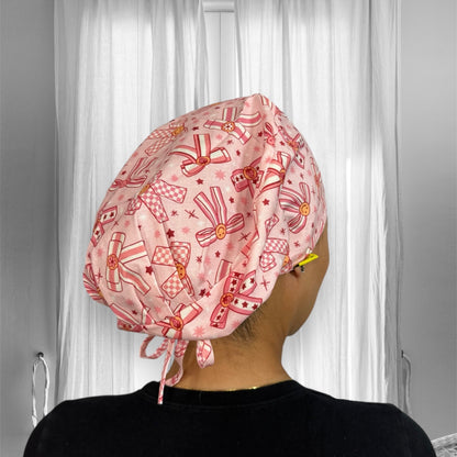 A stylish scrub cap with a playful and colorful design. The cap features a pattern of bows, stars, and buttons in shades of pink and red. The fabric appears soft and the cap is secured at the back with ties.
