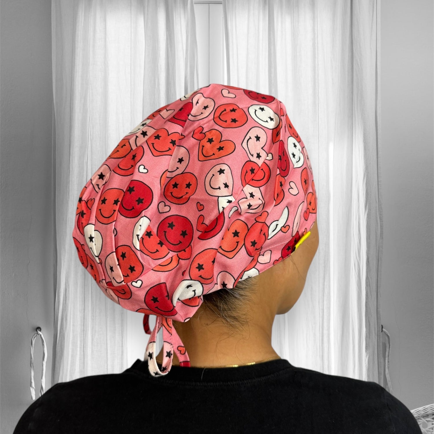 A person wearing a scrub cap with a playful and colorful print. The cap is adorned with various smiley faces and hearts in shades of red, pink, and white.
