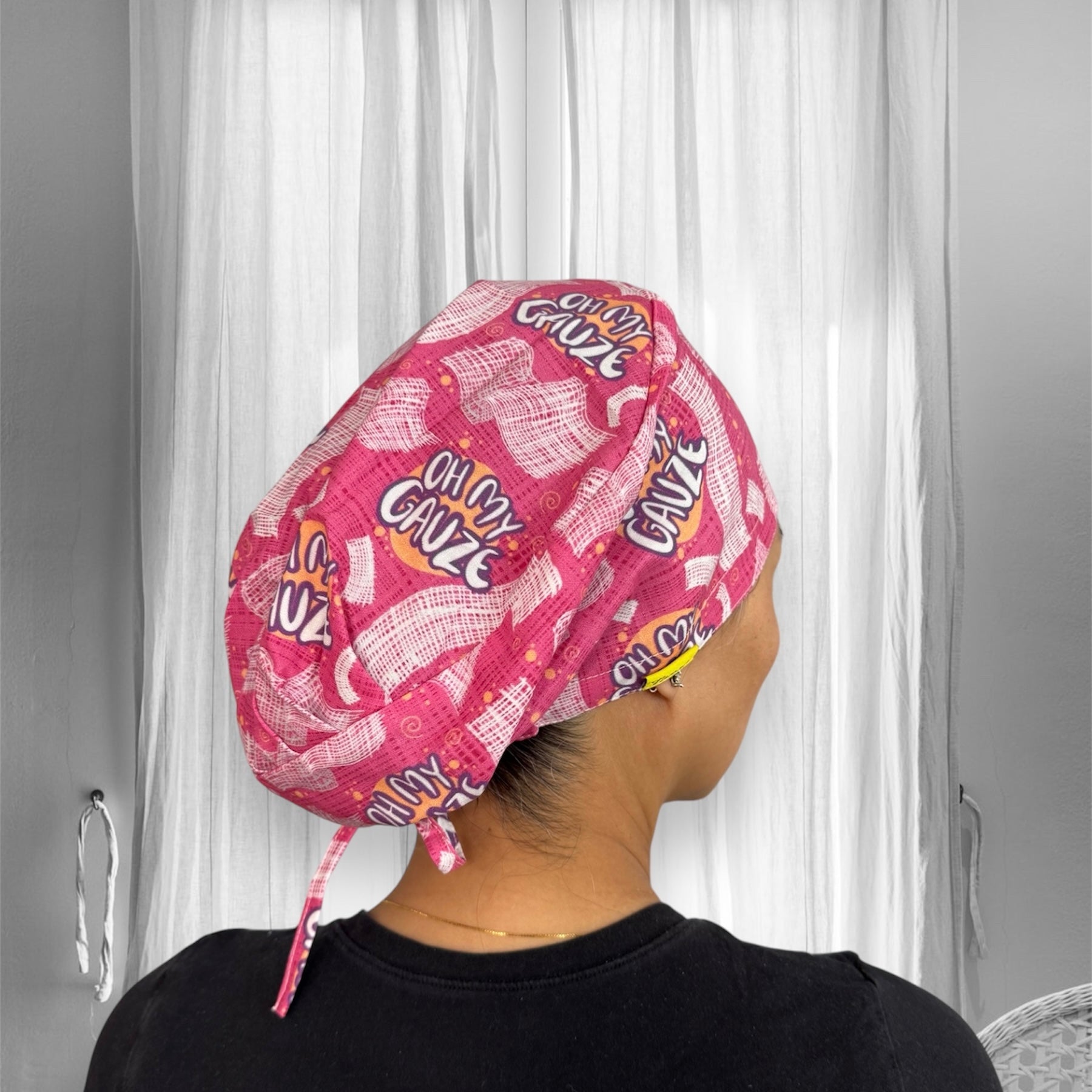 A person wearing a pink scrub cap with playful text design "Oh My Gauze" repeated across the fabric. The cap features a pattern of gauze and abstract shapes.