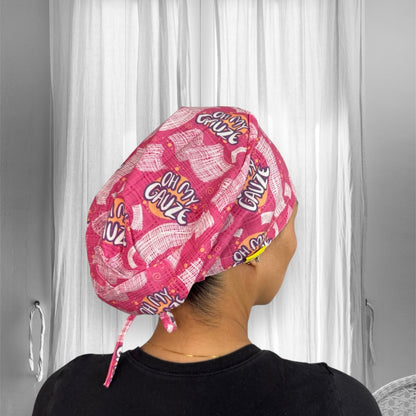 A person wearing a pink scrub cap with playful text design "Oh My Gauze" repeated across the fabric. The cap features a pattern of gauze and abstract shapes.
