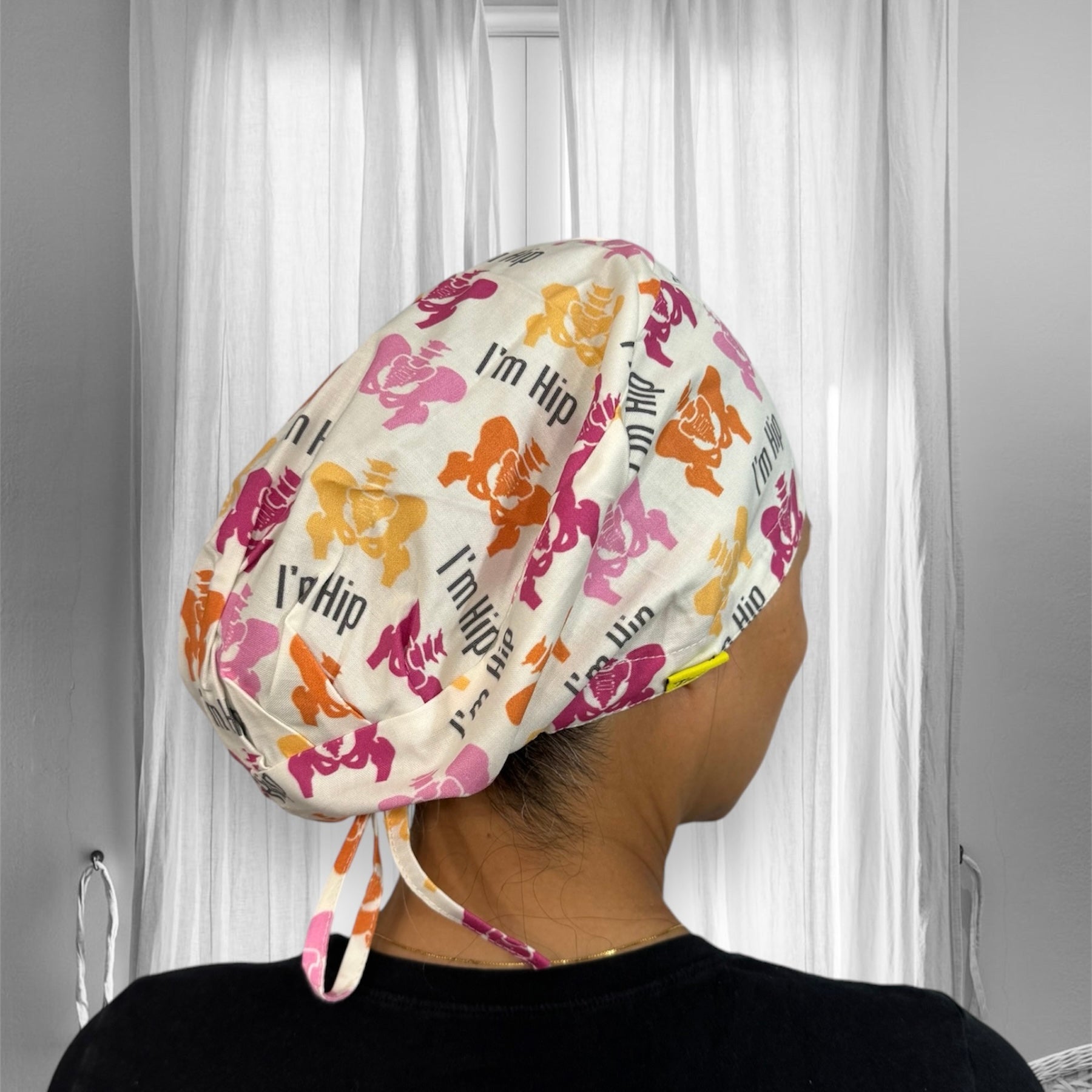 A person wearing a colorful scrub cap with a playful pattern featuring cartoon hippos in pink, orange, and yellow hues. The cap has the phrase "I'm Hip" printed multiple times in a playful font.