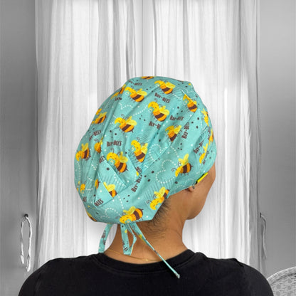 A person is wearing a scrub cap featuring a playful bee pattern. The fabric is light blue with illustrations of smiling bees, each with the word "Bay-Bees" printed next to them.