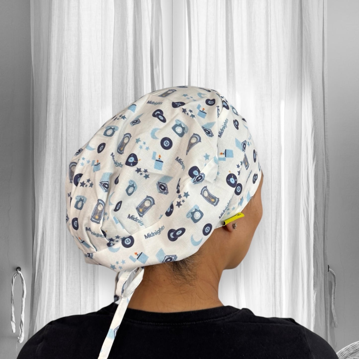 A person wearing a scrub cap. The cap is adorned with a playful pattern featuring various small icons such as clocks, stars, and cameras, along with the word “Midnight” scattered throughout. The fabric has a light background with designs in shades of blue and black. 