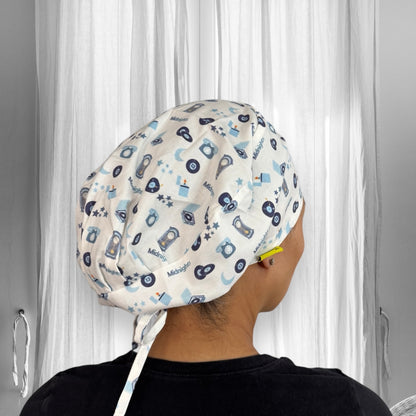 A person wearing a scrub cap. The cap is adorned with a playful pattern featuring various small icons such as clocks, stars, and cameras, along with the word “Midnight” scattered throughout. The fabric has a light background with designs in shades of blue and black. 