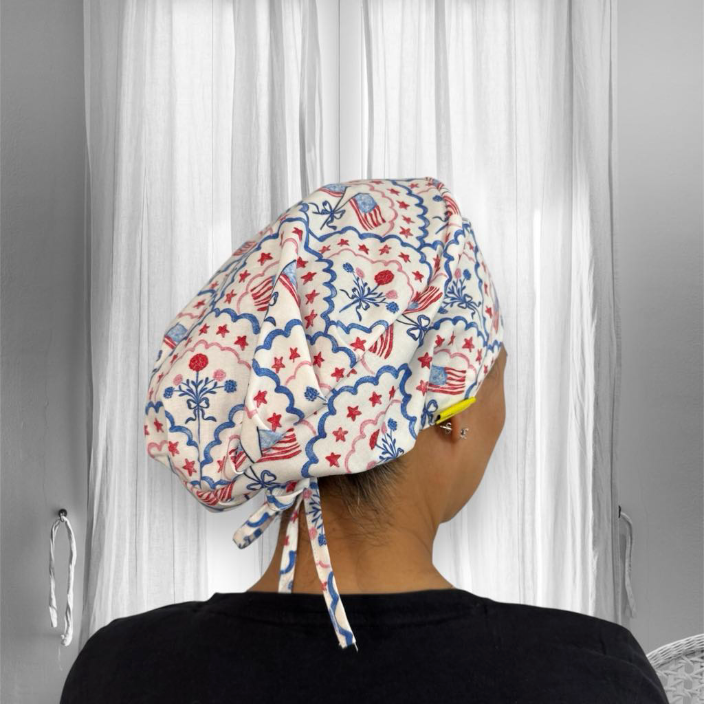 A person wearing a patterned scrub cap. The scrub cap has a festive design featuring red and blue elements, including stars, floral motifs, and stripes, reminiscent of a patriotic or celebratory theme.