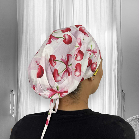 A person wearing a scrub cap made of fabric with a cherry and pink checkerboard pattern. The fabric features vibrant red cherries and pink bows. 