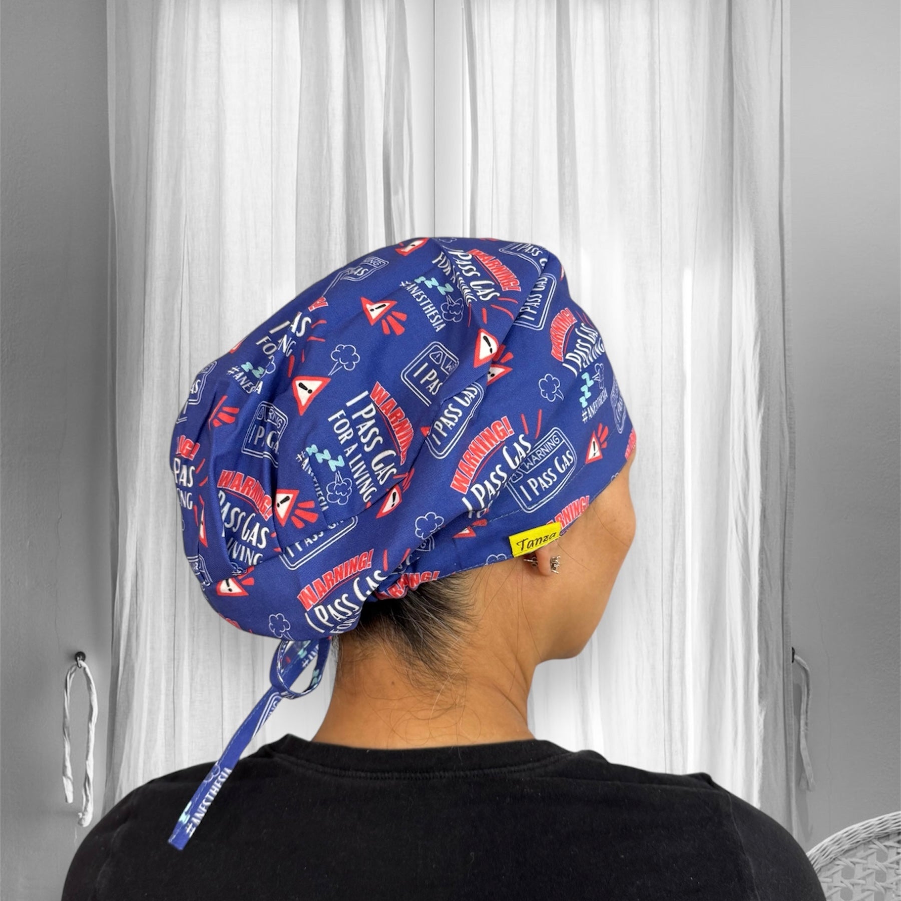 A person wearing a blue scrub cap featuring humorous text and graphics related to "I pass gas" for a living. The cap's design includes warning signs and colorful text.