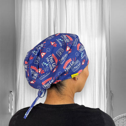 A person wearing a blue scrub cap featuring humorous text and graphics related to "I pass gas" for a living. The cap's design includes warning signs and colorful text.