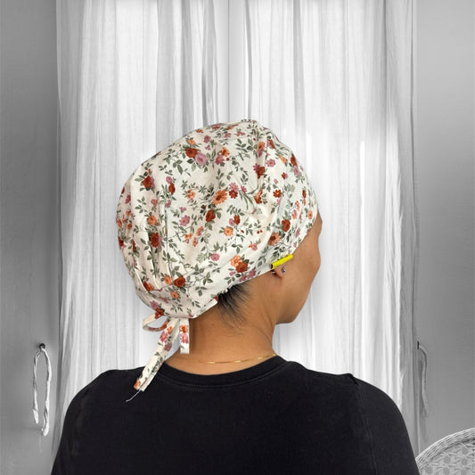 A person wearing a scrub cap with a vibrant floral pattern. The cap is tied at the back, displaying an array of red, orange, and pink flowers with green leaves on a light cream background. 