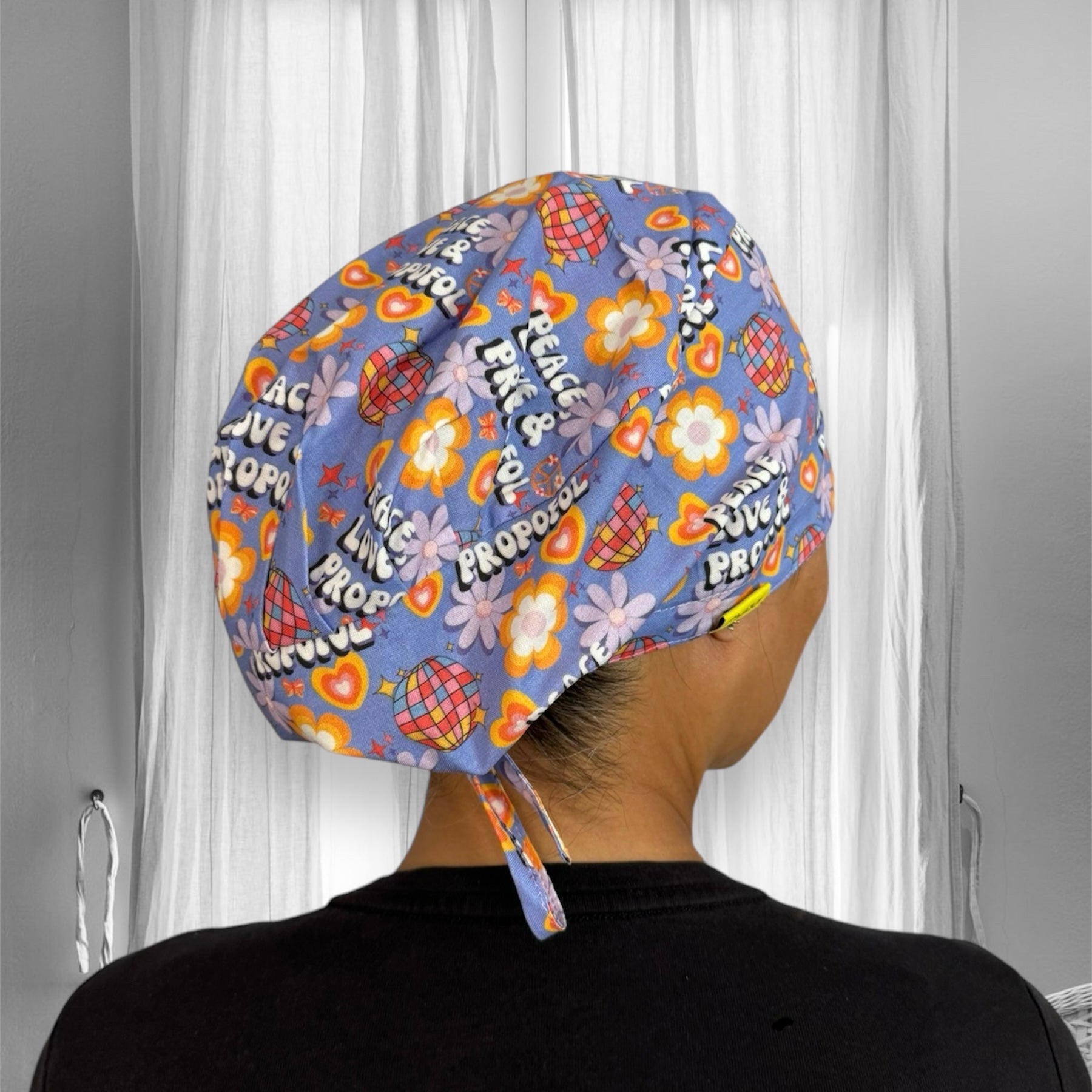 A colorful scrub cap with a vibrant retro design featuring flowers, hearts, disco balls, and text that reads "Peace, Love & Propofol".