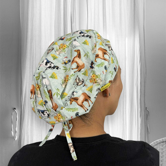 A person wearing a scrub cap with a colorful farm-themed print. The fabric features illustrations of horses, cows, goats, roosters, and floral arrangements on a light pastel green background.