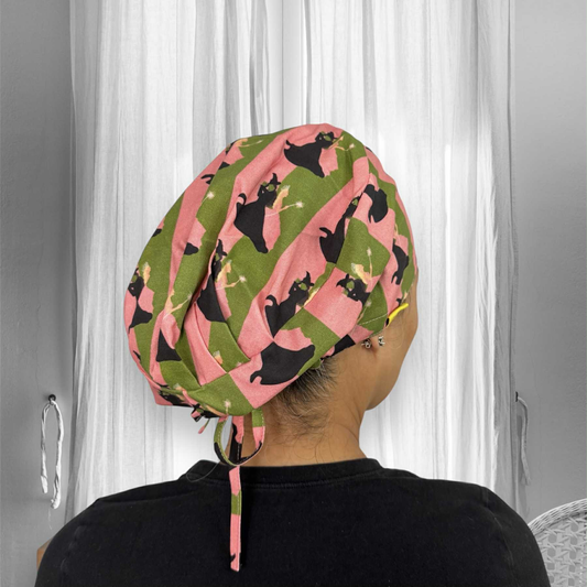 A person wearing a colorful scrub cap. The cap features a playful pattern with alternating pink and green diagonal stripes, adorned with a witch hat illustrations.
