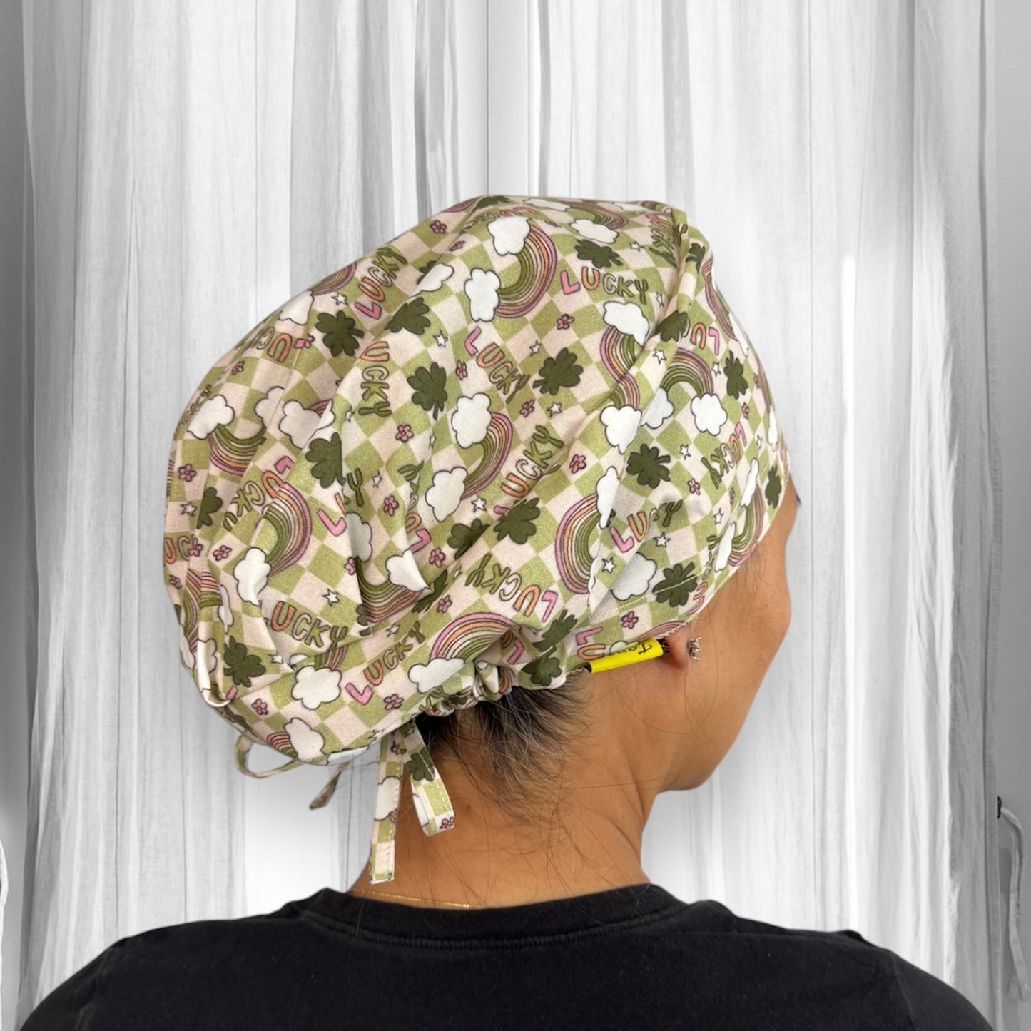 A person  wearing a scrub cap with a pattern of rainbows, clovers, and the word "LUCKY" printed on it. The cap is in shades of green and white, tied at the back.