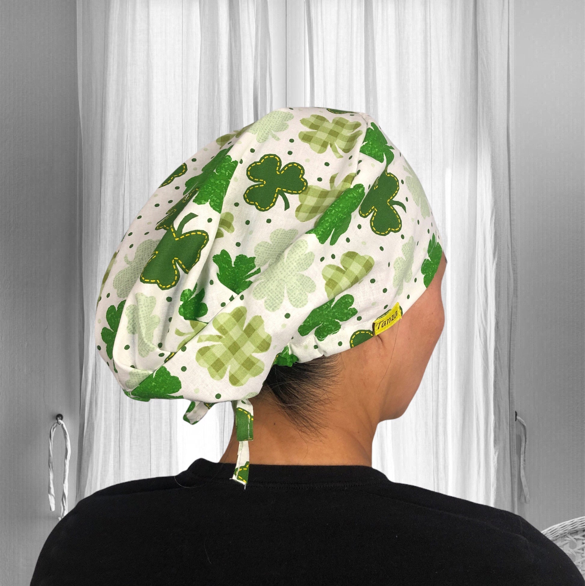 A person wearing a scrub cap with a vibrant green clover pattern. The cap features various shades of green with polka dots and plaid designs, creating a cheerful and festive appearance. 