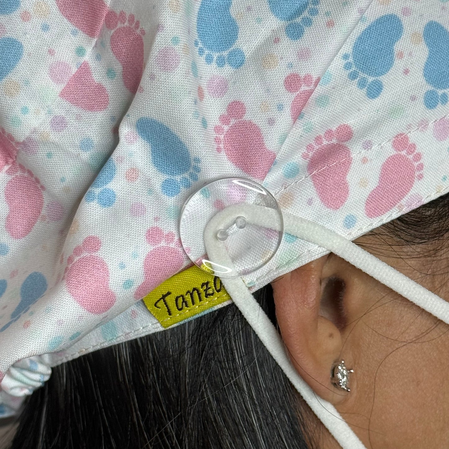 Close-up view of a scrub cap featuring mask loop buttons for added comfort, allowing mask straps to be secured without putting pressure on the ears. The fabric has a playful pattern with pink and blue footprints. Ideal for healthcare professionals who wear masks for extended periods.