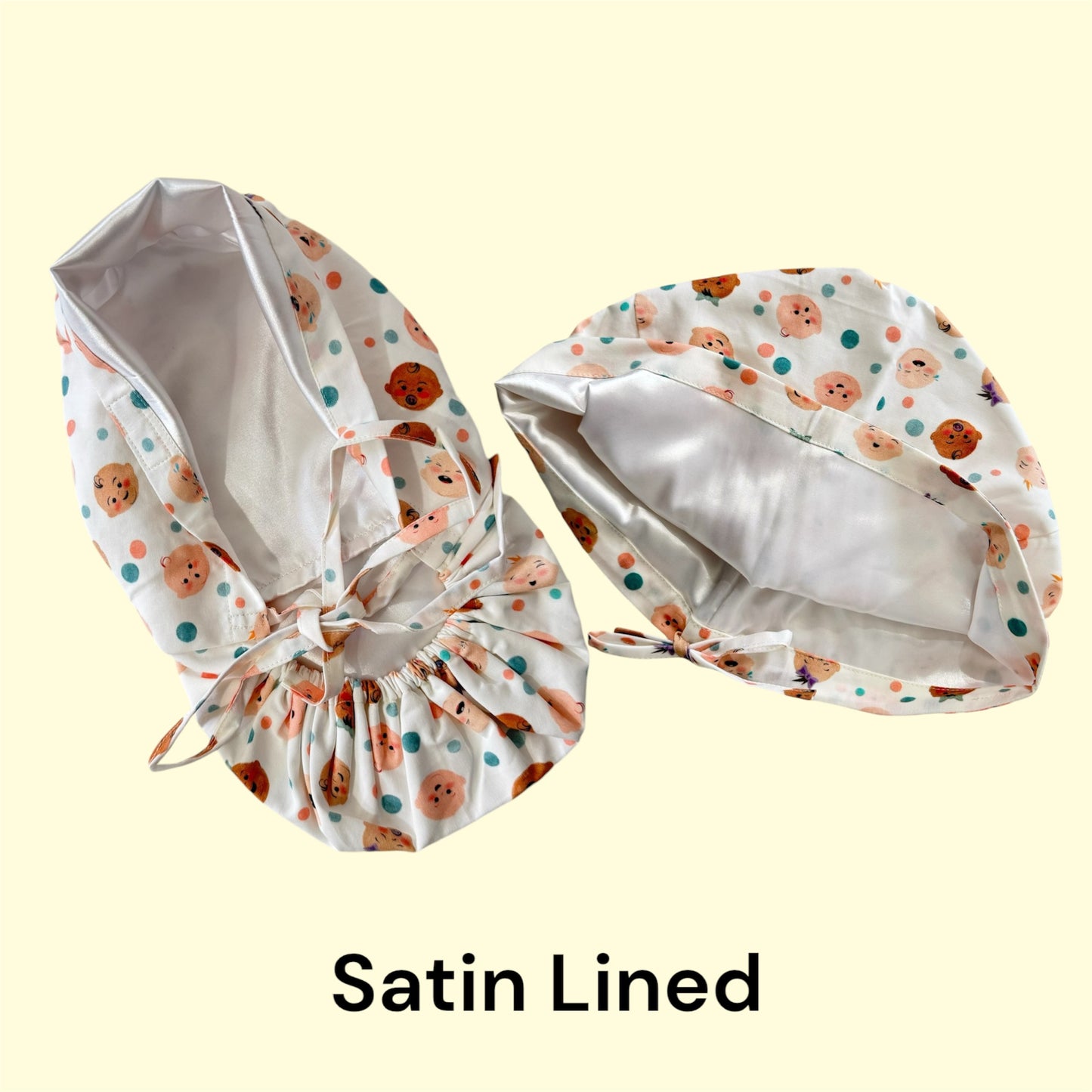 Lay-flat image showcasing a sample of ponytail and Pleated Euro scrub caps with satin lining. The surgical caps feature a smooth satin interior for added comfort and hair protection. The outer fabric displays a playful design with cute baby of diversity faces and pastel colored polka dots. Both scrub caps have adjustable ties at the back for secure and comfortable fit.