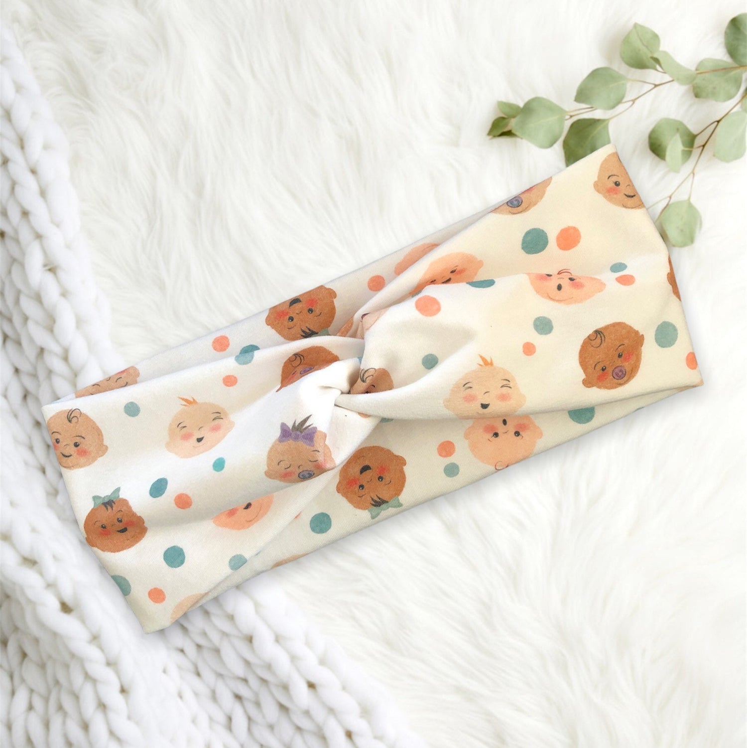 A soft, fabric headband with a playful pattern featuring cute cartoon baby faces and colorful polka dots.