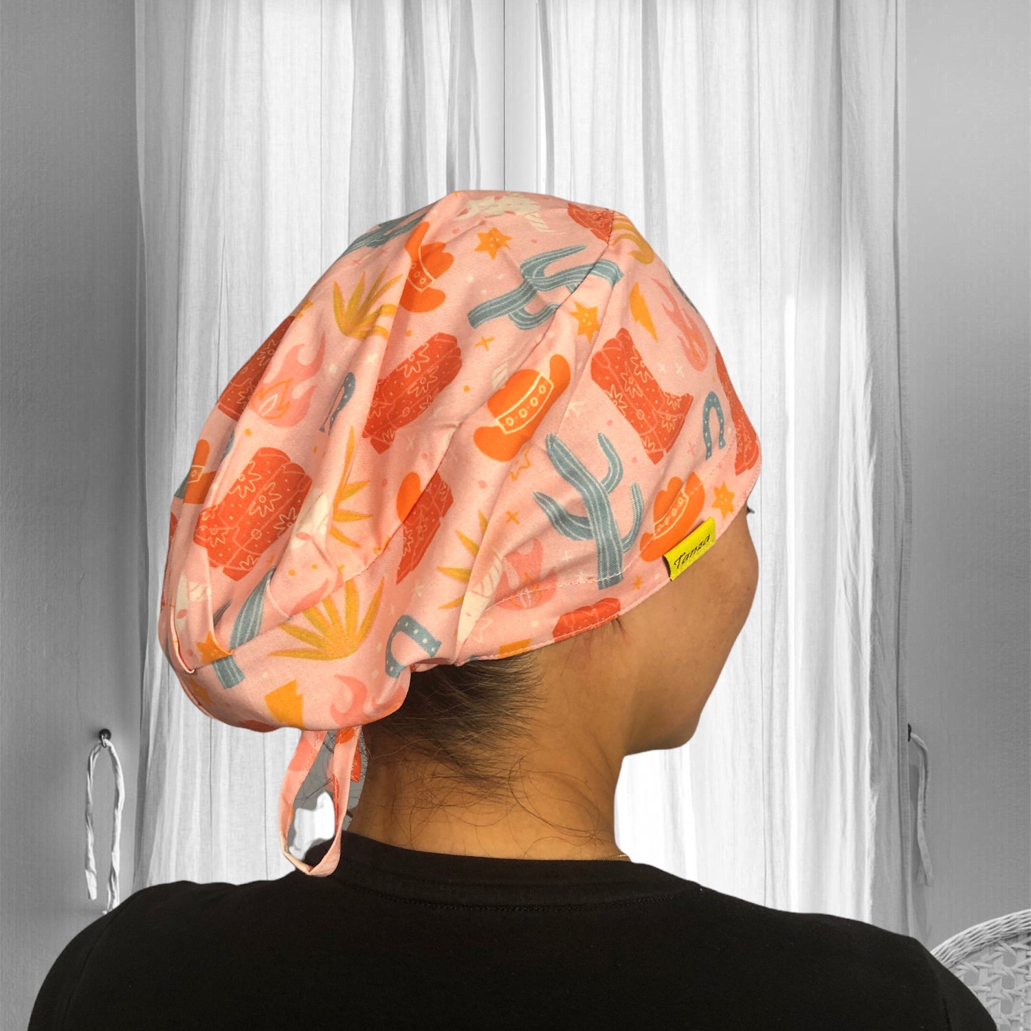 A person wearing a vibrant scrub cap with a playful pattern of cacti, cowboy hats, and horseshoes. The cap is primarily in shades of orange and pink, featuring a light-hearted, Western theme.