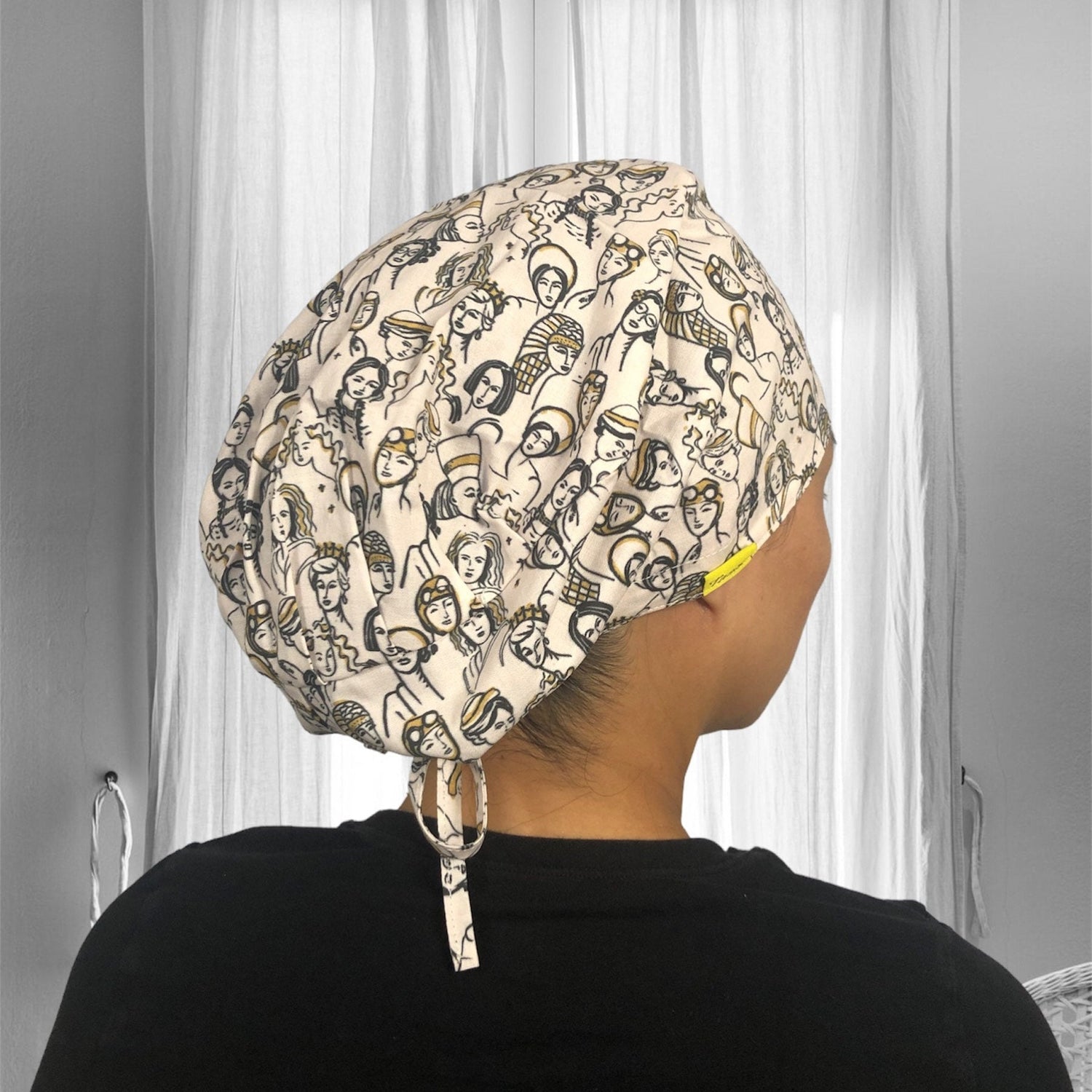 A person wearing a scrub cap adorned with a whimsical pattern of various abstract faces drawn in black and gold on a cream background.