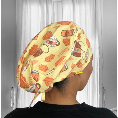 A person is wearing a colorful scrub cap featuring a vibrant autumn-themed pattern. The cap is adorned with illustrations of mugs, leaves, and pumpkins on a light yellow background.