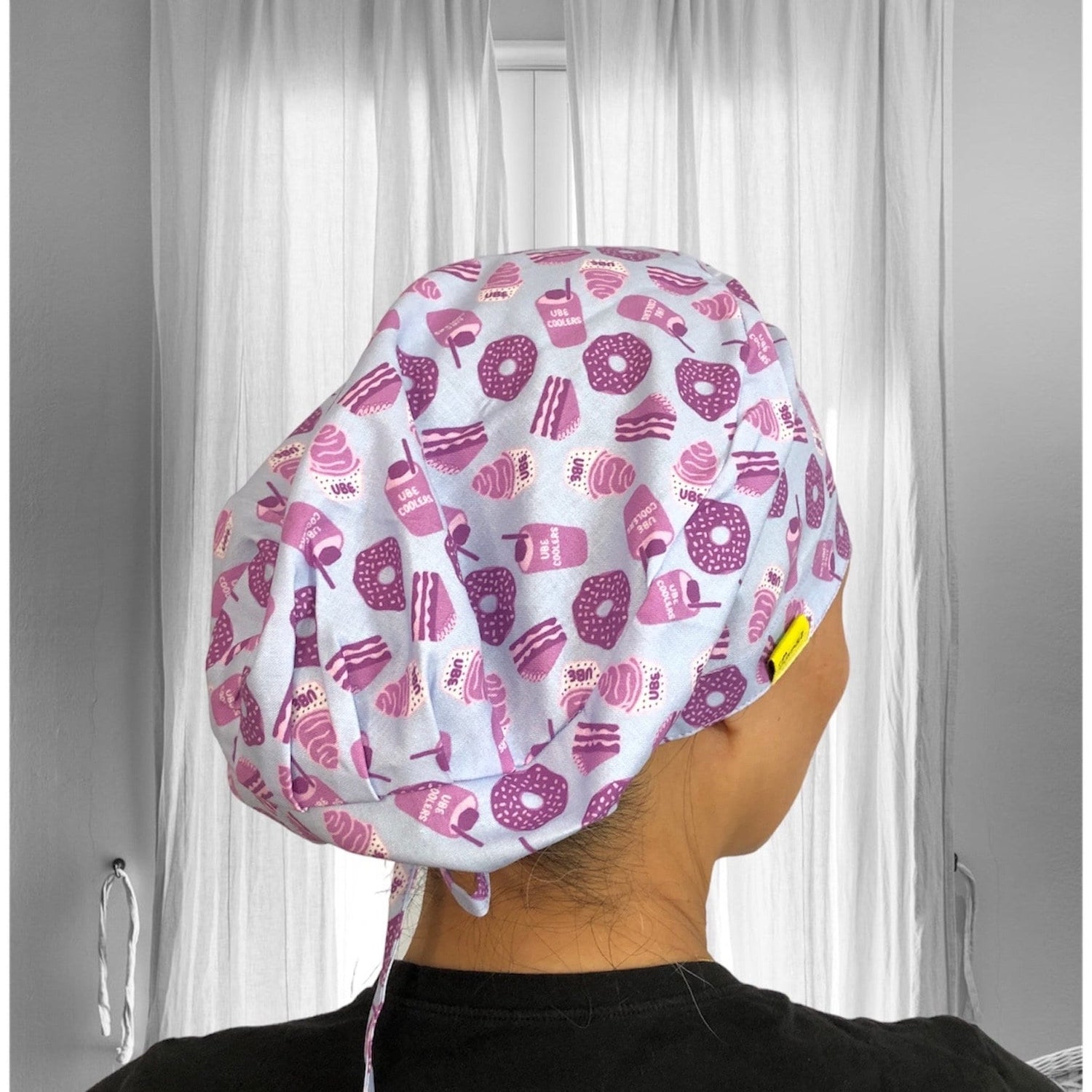 A person wearing a scrub cap with a playful pattern featuring pink and purple doughnuts, ice cream cones, and cakes on a light background.