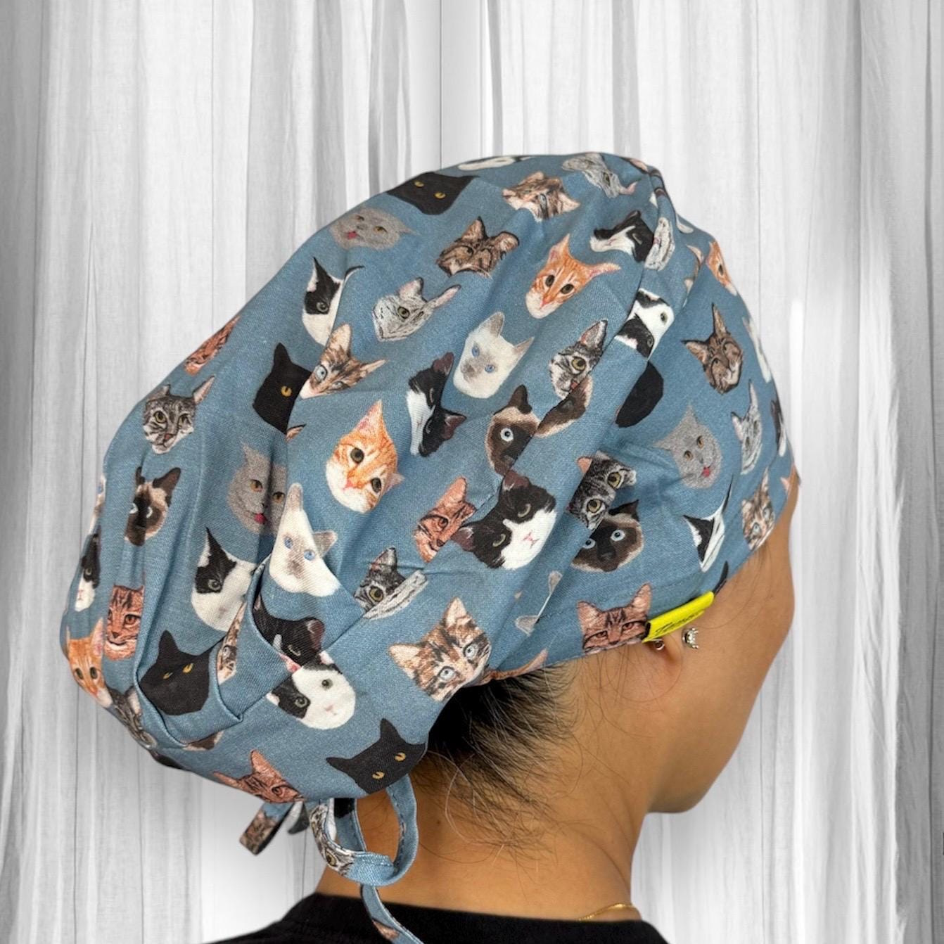 A blue scrub cap adorned with various cat faces, featuring different breeds and expressions. The cap is tied at the back.