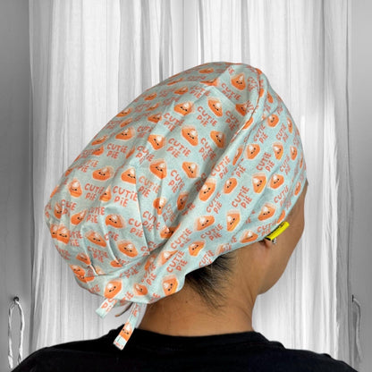 A person wearing a scrub cap with a playful design featuring orange pie graphics and the words "Cutie Pie" repeated across a light blue fabric.
