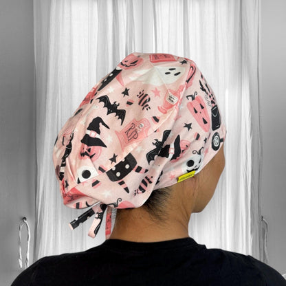 A person wearing a scrub cap with a playful Halloween-themed design. The cap features various festive prints including pumpkins, bats, ghosts, and witches' hats, all in shades of pink, black, and white.
